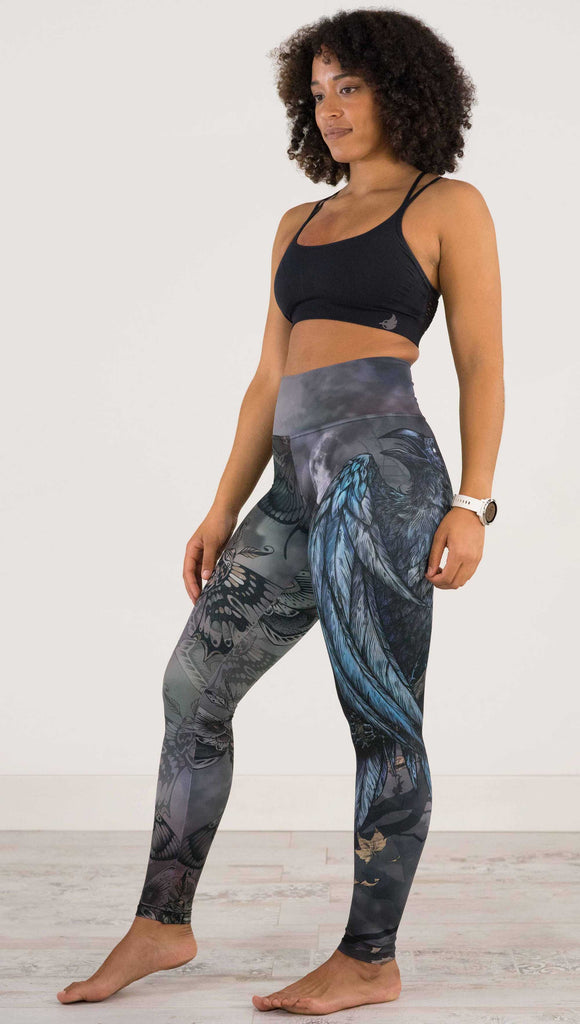 Full body side view of model wearing WERKSHOP Gothica Mashup Leggings. The leggings feature original artwork with moths over a muted/dark oil-slick inspired background on one leg and a spooky scene with a raven and a moon on the other leg.