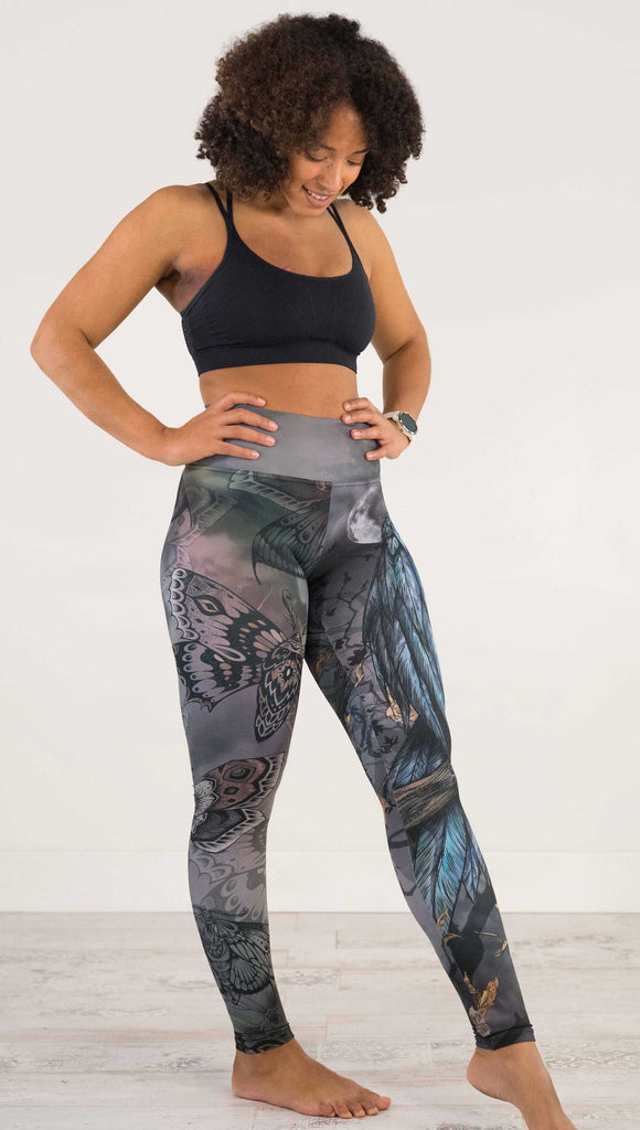 Full body front view of model wearing WERKSHOP Gothica Mashup Leggings. The leggings feature original artwork with moths over a muted/dark oil-slick inspired background on one leg and a spooky scene with a raven and a moon on the other leg.