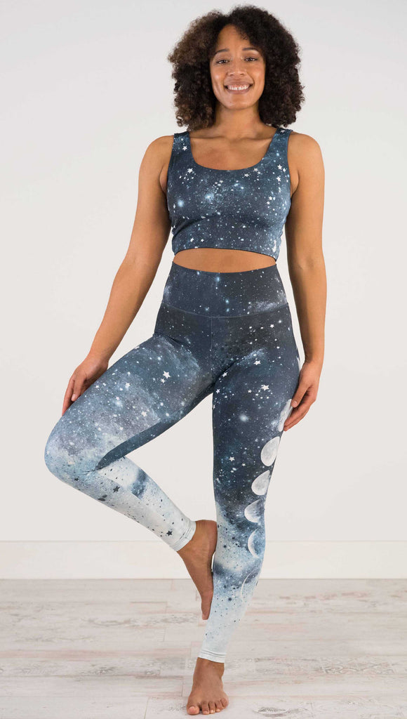 Full body view of a girl wearing our moon phases athleisure leggings - with the phases of the moon printed on the wearer's left leg on a starry night sky with all zodiac constellations in the background