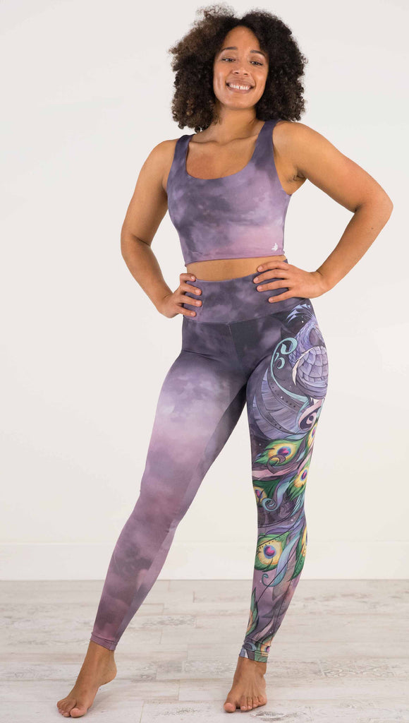 Full body front view of model wearing WERKSHOP Peacock Athleisure Leggings. The artwork on the leggings features a vibrant, colorful peacock down the wearer's left leg over a beautiful warm purple and pink watercolor background.