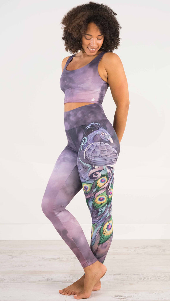 Full body side view of model wearing WERKSHOP Peacock Athleisure Leggings. The artwork on the leggings features a vibrant, colorful peacock down the wearer's left leg over a beautiful warm purple and pink watercolor background.