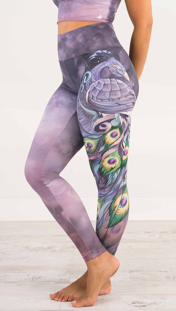 Waist down side view of model wearing WERKSHOP Peacock Athleisure Leggings. The artwork on the leggings features a vibrant, colorful peacock down the wearer's left leg over a beautiful warm purple and pink watercolor background.