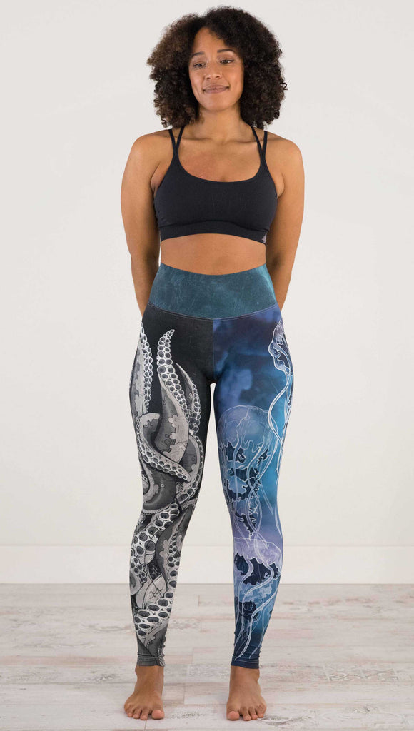 Full body front view of model wearing WERKSHOP Sea Mashup Athleisure Leggings. The artwork on the leggings features hand-drawn black and white tentacles wrapping up the wearers right leg and jellyfish over a blue watercolor background on the left leg. They are tied together with a blue-green under-the-sea themed waistband,