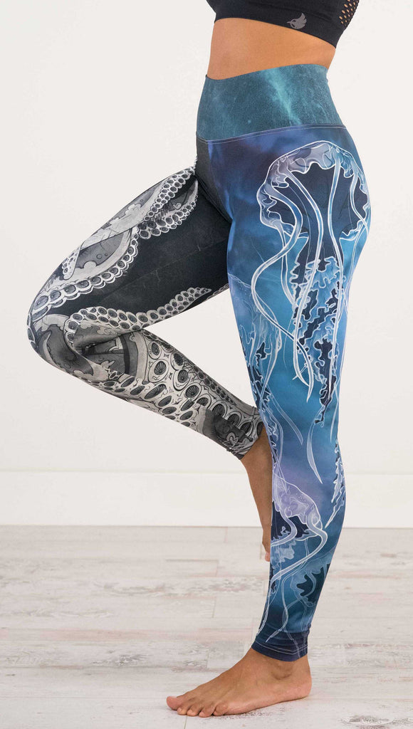 Waist down side view of model wearing WERKSHOP Sea Mashup Athleisure Leggings. The artwork on the leggings features hand-drawn black and white tentacles wrapping up the wearers right leg and jellyfish over a blue watercolor background on the left leg. They are tied together with a blue-green under-the-sea themed waistband,