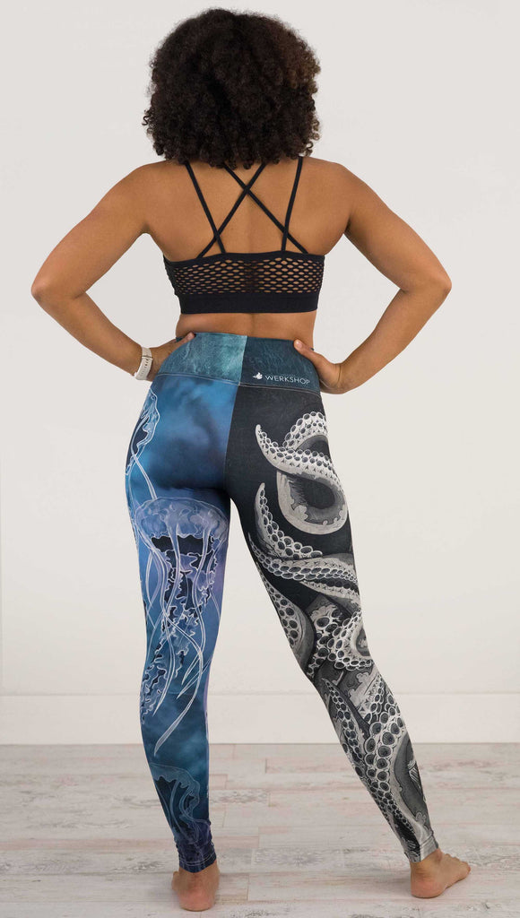 Full body back view of model wearing WERKSHOP Sea Mashup Athleisure Leggings. The artwork on the leggings features hand-drawn black and white tentacles wrapping up the wearers right leg and jellyfish over a blue watercolor background on the left leg. They are tied together with a blue-green under-the-sea themed waistband,