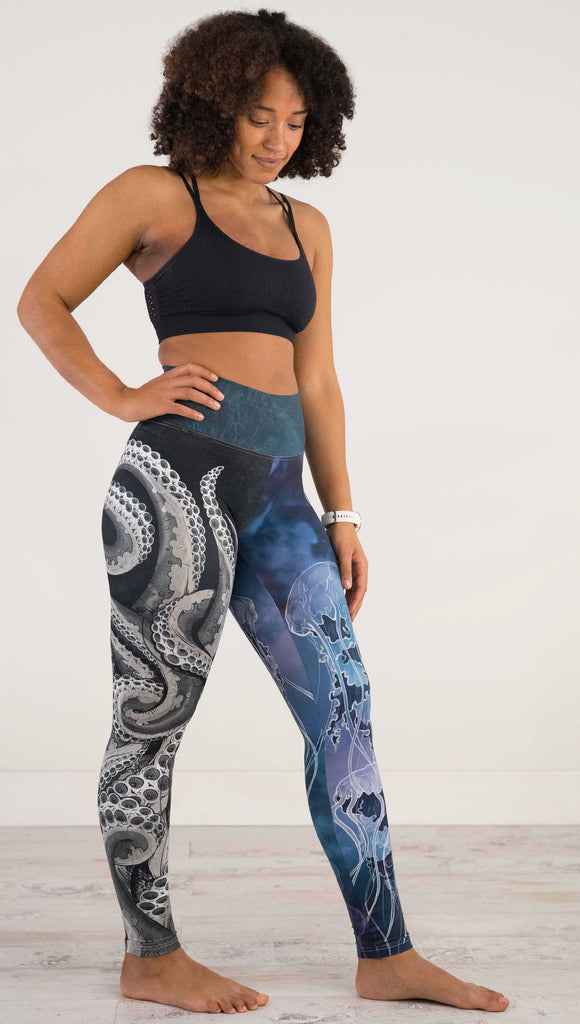 Full body side view of model wearing WERKSHOP Sea Mashup Athleisure Leggings. The artwork on the leggings features hand-drawn black and white tentacles wrapping up the wearers right leg and jellyfish over a blue watercolor background on the left leg. They are tied together with a blue-green under-the-sea themed waistband,