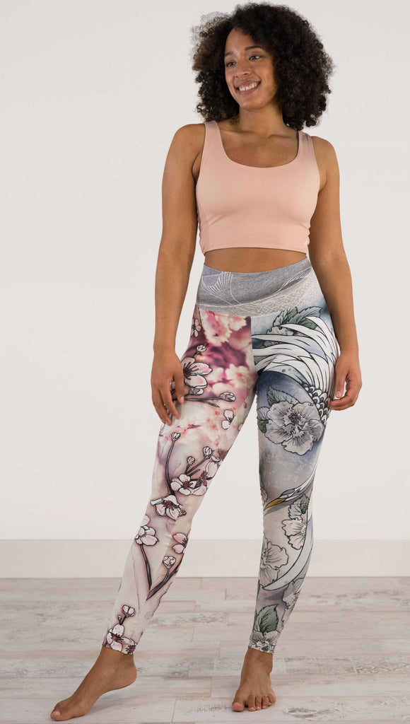 Full body front view of model wearing WERKSHOP Zen Mashup Athleisure Leggings. The leggigns are printed with pink cherry blossoms on the wearer's left leg and a graceful swooping crane on the opposite leg. There is also an abstract outline drawing of a koi fish on the waistband.
