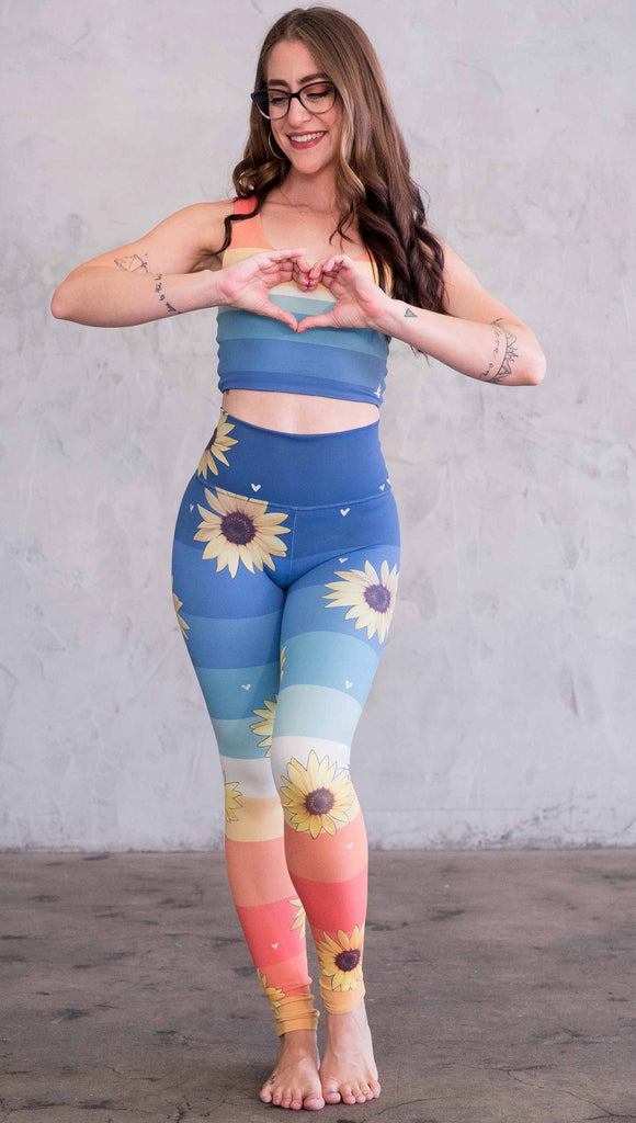 Full body front view of model wearing WERKSHOP Sunflower Athleisure leggings. The leggings have wide horizontal stripes with dark blue at the waistband, to aqua and pale green at the mid thigh leading to cream at the knee and orange and red tones to the ankle. There are large photo-real sunflowers and tiny hand sketched off-white hearts sprinkled throughout.