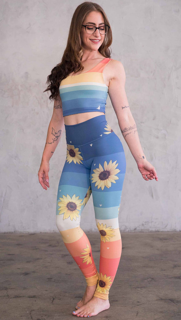 Full body front view of model wearing WERKSHOP Sunflower Athleisure leggings. The leggings have wide horizontal stripes with dark blue at the waistband, to aqua and pale green at the mid thigh leading to cream at the knee and orange and red tones to the ankle. There are large photo-real sunflowers and tiny hand sketched off-white hearts sprinkled throughout.