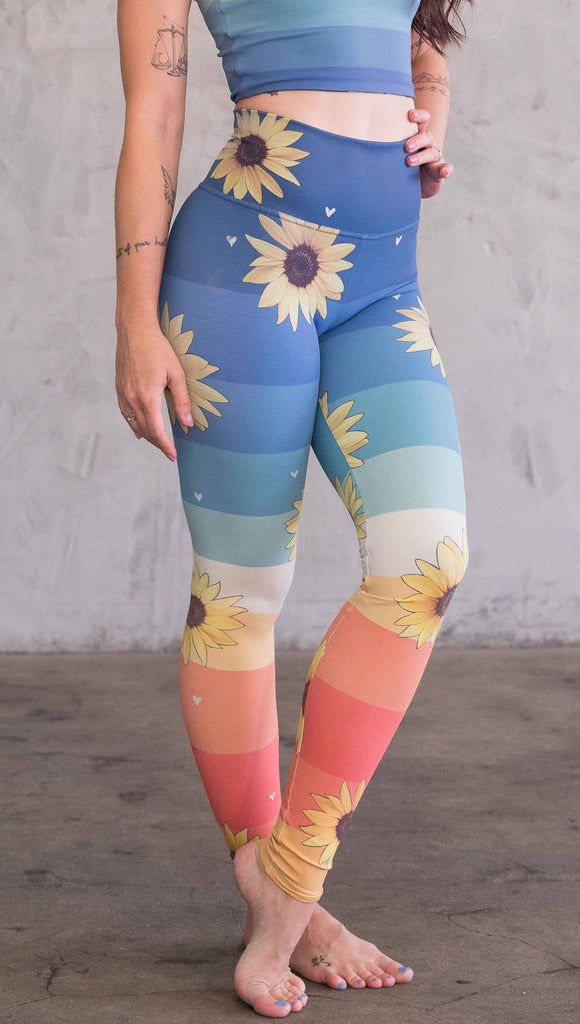 Waist down front view of model wearing WERKSHOP Sunflower Athleisure leggings. The leggings have wide horizontal stripes with dark blue at the waistband, to aqua and pale green at the mid thigh leading to cream at the knee and orange and red tones to the ankle. There are large photo-real sunflowers and tiny hand sketched off-white hearts sprinkled throughout.