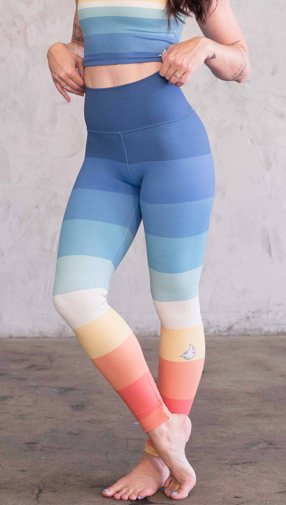 Waist down front view of model wearing WERKSHOP Vintage Rainbow Athleisure leggings. The leggings have wide horizontal stripes with dark blue at the waistband, to auqua and pale green at the mid thigh leading to cream at the knee and orange and red tones to the ankle.