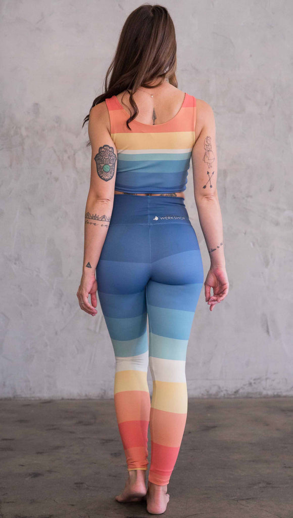 Full body back view of model wearing WERKSHOP Vintage Rainbow Athleisure leggings. The leggings have wide horizontal stripes with dark blue at the waistband, to auqua and pale green at the mid thigh leading to cream at the knee and orange and red tones to the ankle.