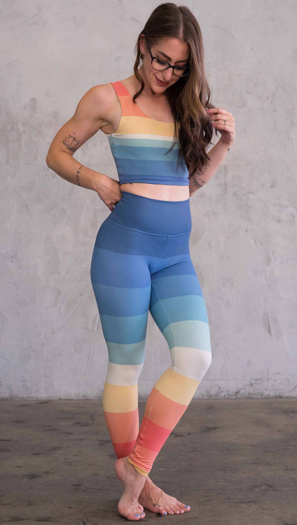 Full body side view of model wearing WERKSHOP Vintage Rainbow Athleisure leggings. The leggings have wide horizontal stripes with dark blue at the waistband, to auqua and pale green at the mid thigh leading to cream at the knee and orange and red tones to the ankle.