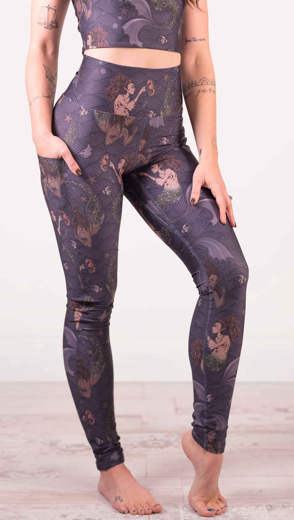 Waist down front view of model wearing WERKSHOP featherlight leggings with Original Mermaids artwork. The mermaids have small intricate details on the fins and are swimming with seahorses and angel fish over a dark blue background with waves. The leggings have large pockets on each hip large enough to hold a phone.