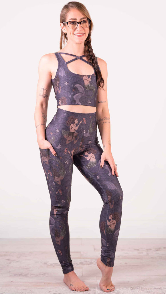 Full body front view of model wearing WERKSHOP featherlight leggings with Original Mermaids artwork. The mermaids have small intricate details on the fins and are swimming with seahorses and angel fish over a dark blue background with waves. The leggings have large pockets on each hip large enough to hold a phone.