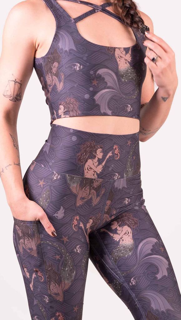 Zoomed in view of model wearing WERKSHOP featherlight leggings with Original Mermaids artwork. The mermaids have small intricate details on the fins and are swimming with seahorses and angel fish over a dark blue background with waves. The leggings have large pockets on each hip large enough to hold a phone.