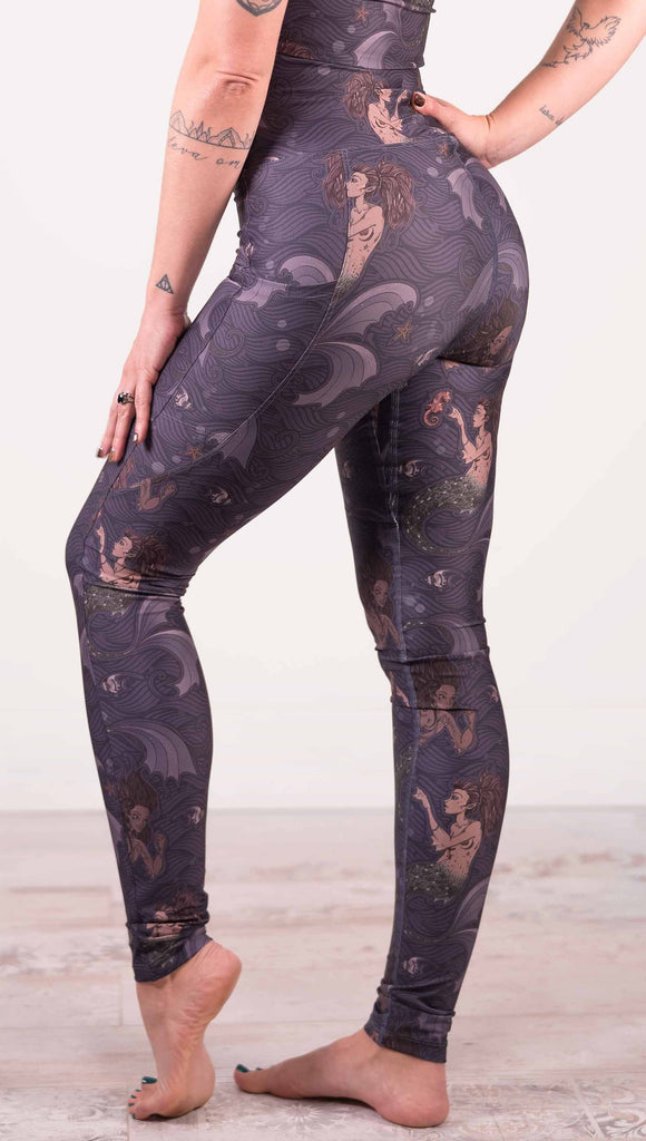 Waist down back view of model wearing WERKSHOP featherlight leggings with Original Mermaids artwork. The mermaids have small intricate details on the fins and are swimming with seahorses and angel fish over a dark blue background with waves. The leggings have large pockets on each hip large enough to hold a phone.