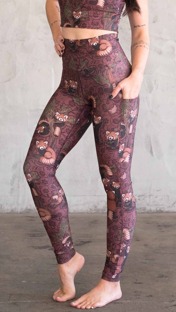 Side view of model wearing WERKSHOP Red Panda Leggings. The artwork is dark red with clusters of cute red pandas playing on trees. The leggings have phone pockets on both legs.