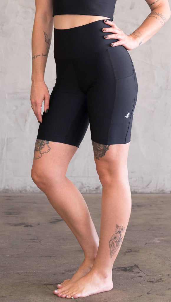 Waist down view of model wearing WERKSHOP bicycle length shorts in featherlight fabric. Black color with side cell phone pockets and a small eagle logo on the wearers left side