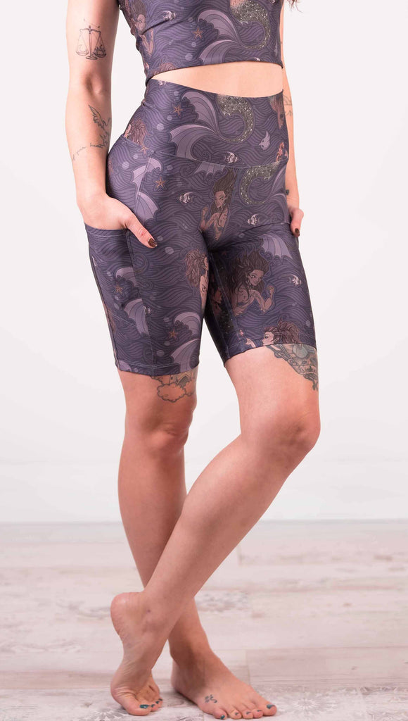 Waist down front view of model wearing WERKSHOP featherlight bicycle length shorts with Original Mermaids artwork. The mermaids have small intricate details on the fins and are swimming with seahorses and angel fish over a dark blue background with waves. The shorts have large pockets on each hip large enough to hold a phone.
