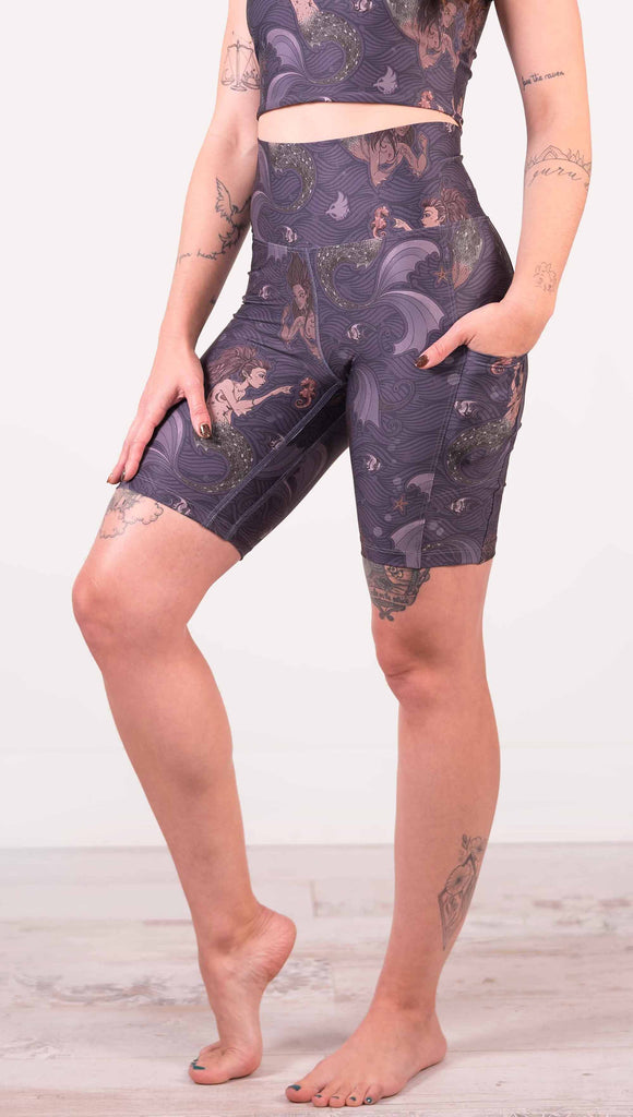 Waist down front view of model wearing WERKSHOP featherlight bicycle length shorts with Original Mermaids artwork. The mermaids have small intricate details on the fins and are swimming with seahorses and angel fish over a dark blue background with waves. The shorts have large pockets on each hip large enough to hold a phone.