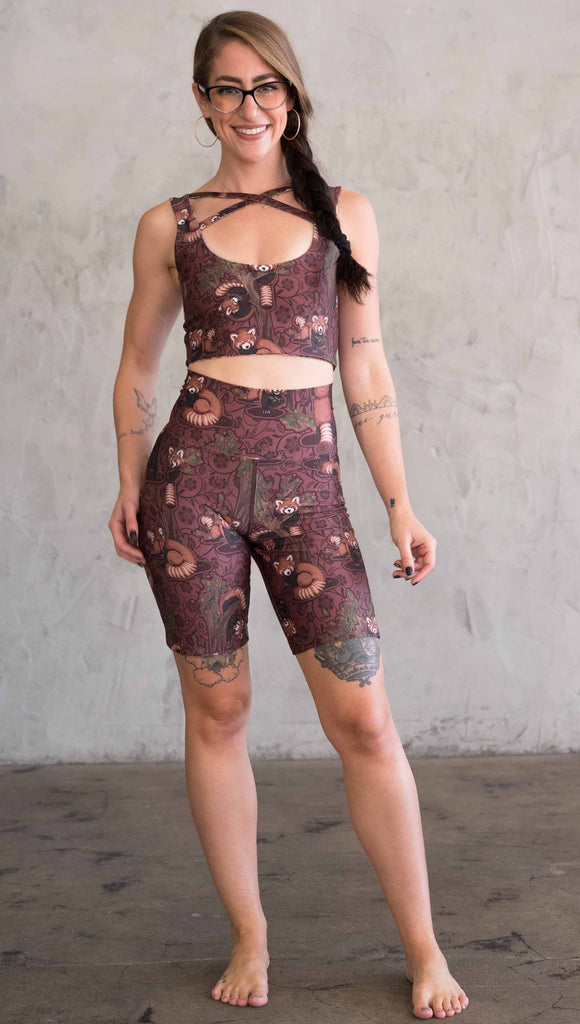 Full body front view of model wearing WERKSHOP Red Panda Bicycle Length shorts. The artwork on the shorts features adorable little red pandas playing an having snacks next to abstract trees on a burgundy background.