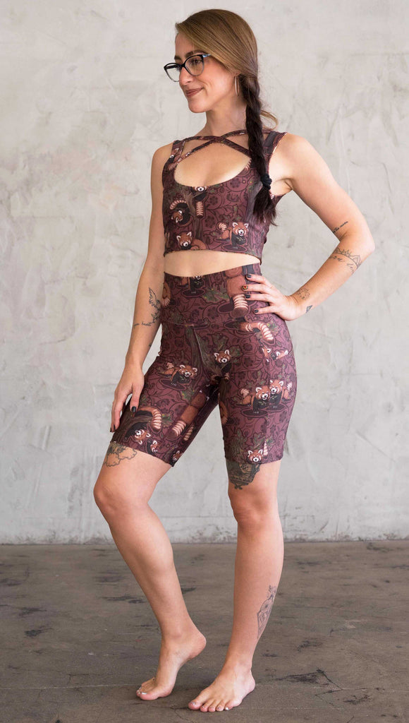 Full body side view of model wearing WERKSHOP Red Panda Bicycle Length shorts. The artwork on the shorts features adorable little red pandas playing an having snacks next to abstract trees on a burgundy background.