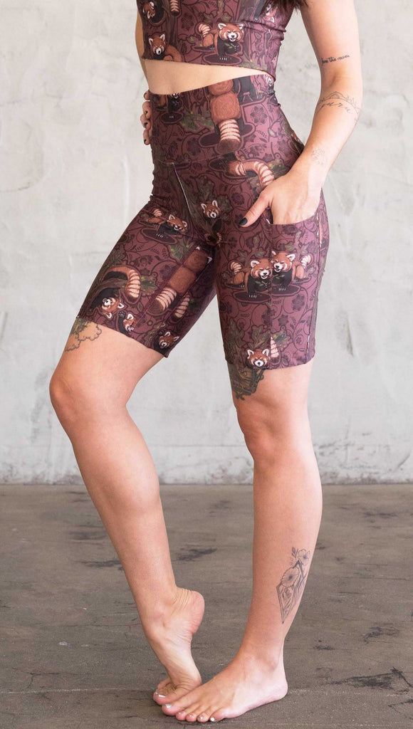 Waist down side view of model wearing WERKSHOP Red Panda Bicycle Length shorts. The artwork on the shorts features adorable little red pandas playing an having snacks next to abstract trees on a burgundy background.