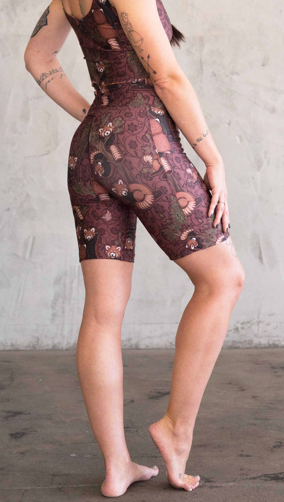 Waist down back view of model wearing WERKSHOP Red Panda Bicycle Length shorts. The artwork on the shorts features adorable little red pandas playing an having snacks next to abstract trees on a burgundy background.