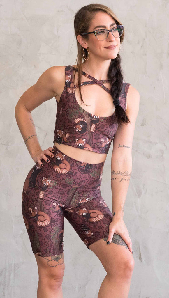 Mid-body zoomed-in view of model wearing WERKSHOP Red Panda Bicycle Length shorts. The artwork on the shorts features adorable little red pandas playing an having snacks next to abstract trees on a burgundy background.