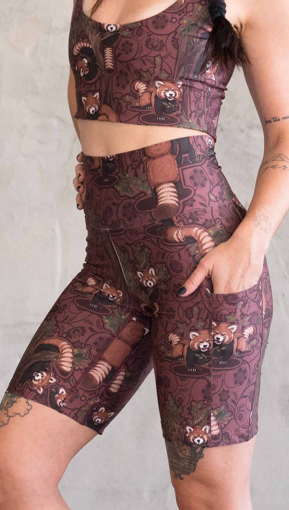 Zoomed in view of model wearing WERKSHOP Red Panda Bicycle Length shorts. The artwork on the shorts features adorable little red pandas playing an having snacks next to abstract trees on a burgundy background.