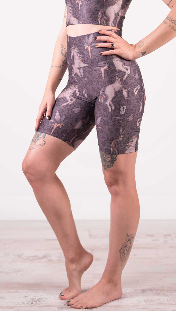 Wait down front view of model wearing WERKSHOP featherlight bicycle length shorts with Original Unicorn artwork. The unicorns have soft rainbow colored hair and a small pixie friend over a purple background. The shorts have large pockets on each hip large enough to hold a phone.