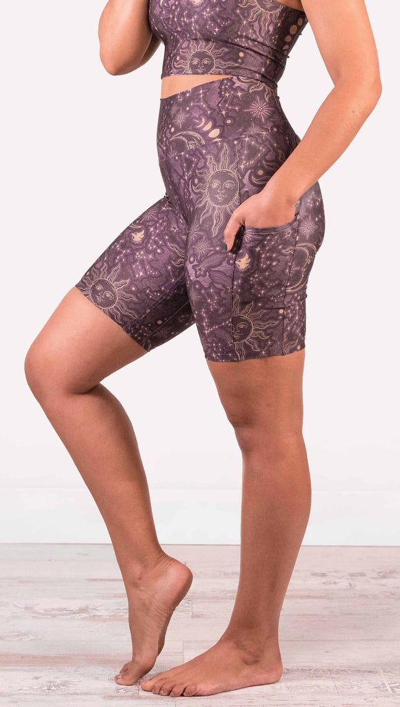 Model wearing WERKSHOP Zodiac Bicycle Length Shorts. They are high waist and feature pockets on both legs. The zodiac themed artwork shows a hand-drawn sun and moon with the moon phases, shooting stars and all 12 zodiac constellations in gold over a dark purple background. The length of the shorts hit in the mid thigh, above the knee.