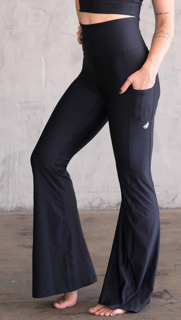 Waist down view of model wearing WERKSHOP Featherlight Bell Bottoms in solid black. The bells have a small reflective eagle logo on the wearer's left side pocket.