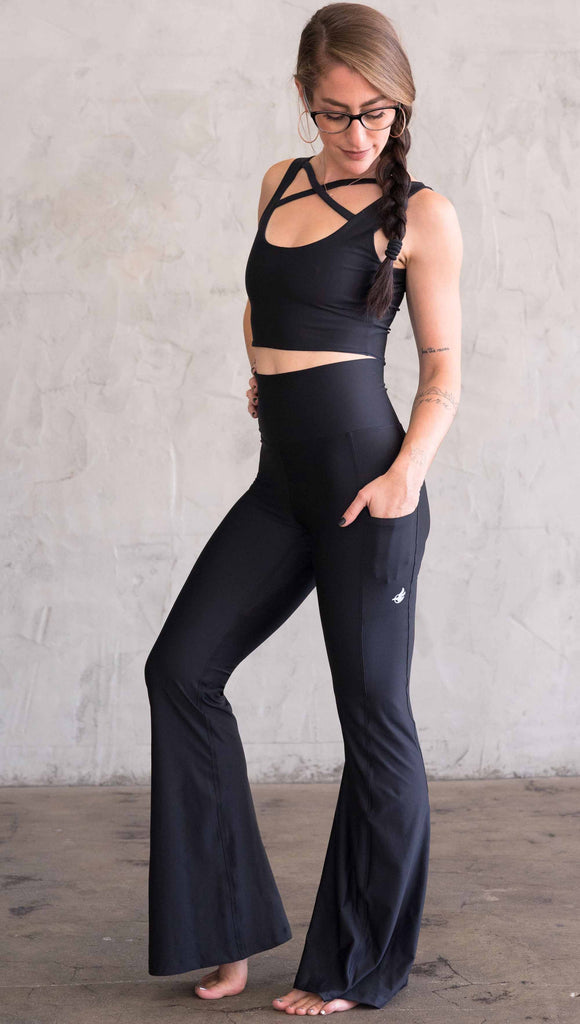 Full body side view of model wearing WERKSHOP Featherlight Bell Bottoms in solid black. The bells have a small reflective eagle logo on the wearer's left side pocket.
