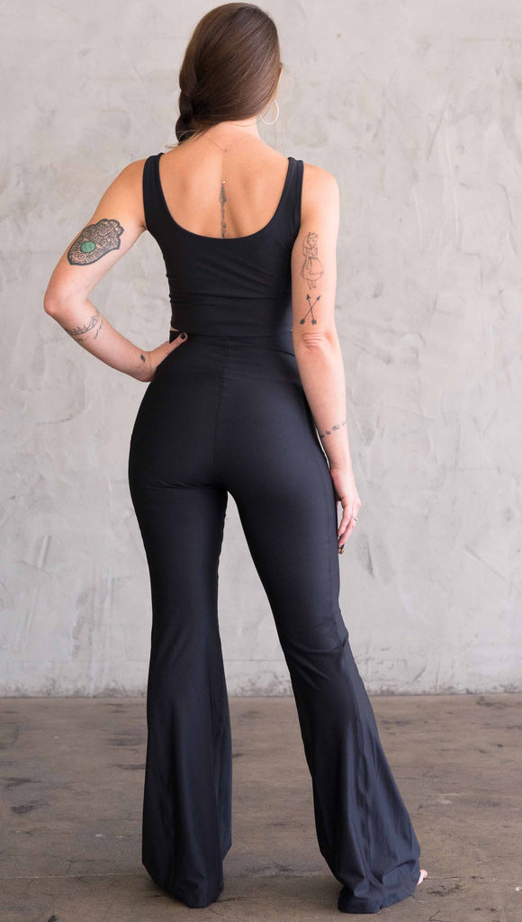 Full body back view of model wearing WERKSHOP Featherlight Bell Bottoms in solid black. The bells have a small reflective eagle logo on the wearer's left side pocket.