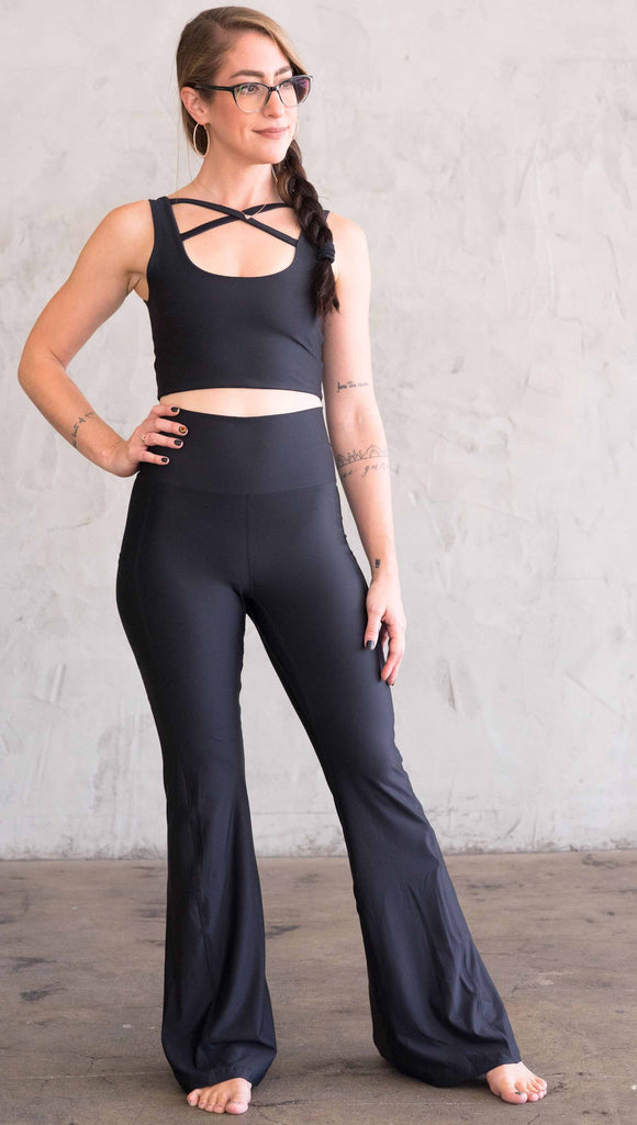 Full body front view of model wearing WERKSHOP Featherlight Bell Bottoms in solid black. The bells have a small reflective eagle logo on the wearer's left side pocket.