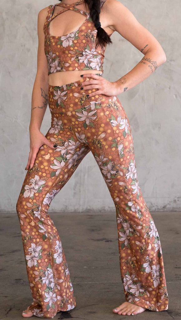 Front view of model wearing WERKSHOP Honeybee Bell Bottoms. The leggings have clusters of honeybees and flowers and phone pockets on each leg