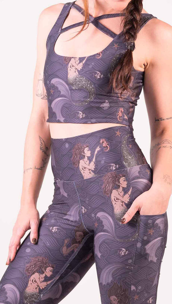 Zoomed in view of model wearing WERKSHOP featherlight bell bottom flares shorts with Original Mermaids artwork. The mermaids have small intricate details on the fins and are swimming with seahorses and angel fish over a dark blue background with waves. The pants have large pockets on each hip large enough to hold a phone.
