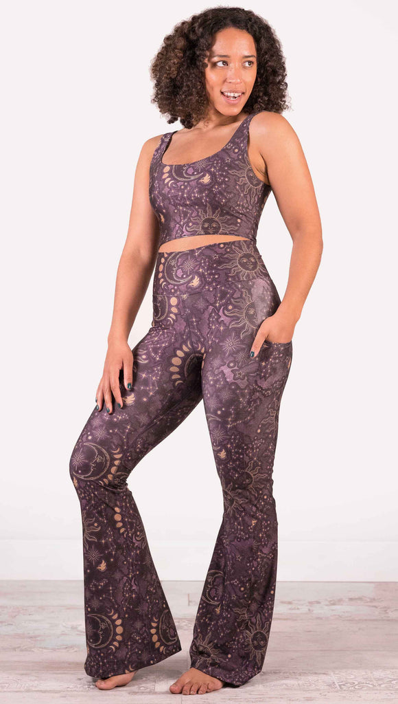Model wearing WERKSHOP Zodiac Bell Bottoms. They are high waist and feature pockets on both legs. The zodiac themed artwork shows a hand-drawn sun and moon with the moon phases, shooting stars and all 12 zodiac constellations in gold over a dark purple background.