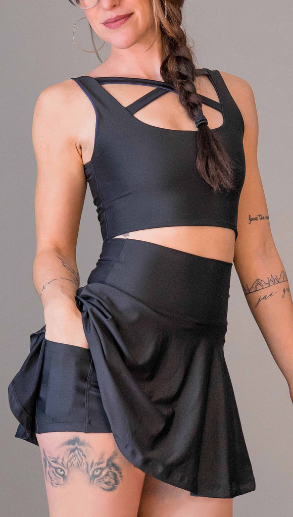 Zoomed in view of model wearing WERKSHOP Tennis Skirt, with the skirt lifted up to show the shorts and pockets underneath. In solid black color
