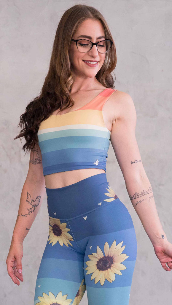 Front view of model wearing WERKSHOP Sunflower Rainbow Top. The top has vintage colored rainbow stripes on both sides. The stripes start orange at the shoulders, yellow and cream across the chest and then blue at the sweep. One side also has sunflowers and small cream colored hearts.