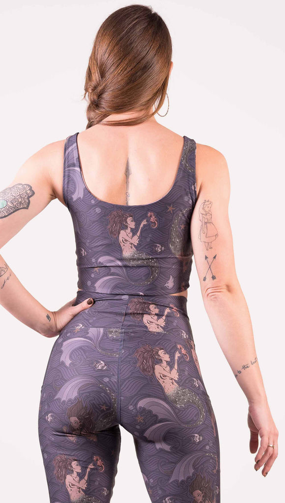 Back view of model wearing WERKSHOP Four-Way Reversible Top with Original Mermaids artwork on one side and Unicorn artwork on the opposite side. She is shown wearing the mermaids on the outside with the “X” strap detail in the front. The mermaids have small intricate details on the fins and are swimming with seahorses over a dark blue background.