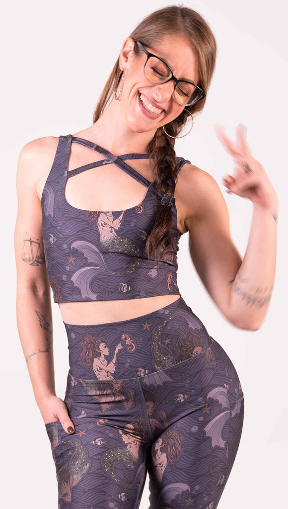 Front view of model wearing WERKSHOP Four-Way Reversible Top with Original Mermaids artwork on one side and Unicorn artwork on the opposite side. She is shown wearing the mermaids on the outside with the “X” strap detail in the front. The mermaids have small intricate details on the fins and are swimming with seahorses over a dark blue background.