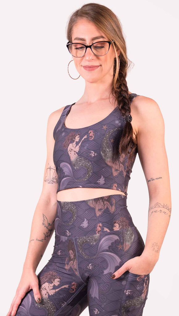 Front view of model wearing WERKSHOP Four-Way Reversible Top with Original Mermaids artwork on one side and Unicorn artwork on the opposite side. She is shown wearing the mermaids on the outside with the “X” strap detail in the back. The mermaids have small intricate details on the fins and are swimming with seahorses over a dark blue background.