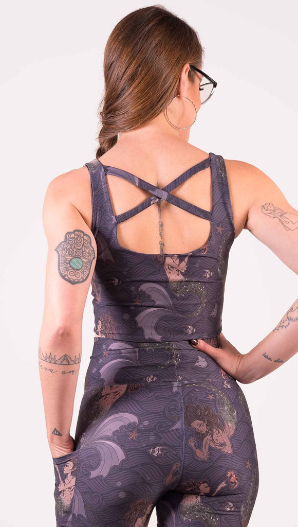 Back view of model wearing WERKSHOP Four-Way Reversible Top with Original Mermaids artwork on one side and Unicorn artwork on the opposite side. She is shown wearing the mermaids on the outside with the “X” strap detail in the back. The mermaids have small intricate details on the fins and are swimming with seahorses over a dark blue background.