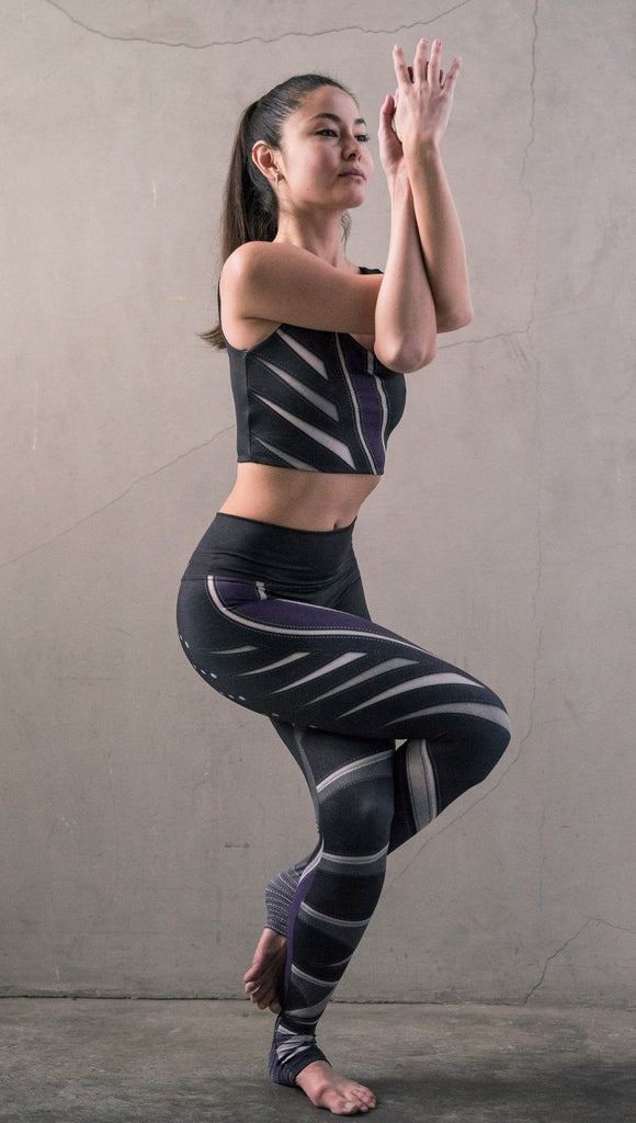 Model in standing yoga pose wearing black printed full-length leggings with purple and gray stripe design and matching sports top