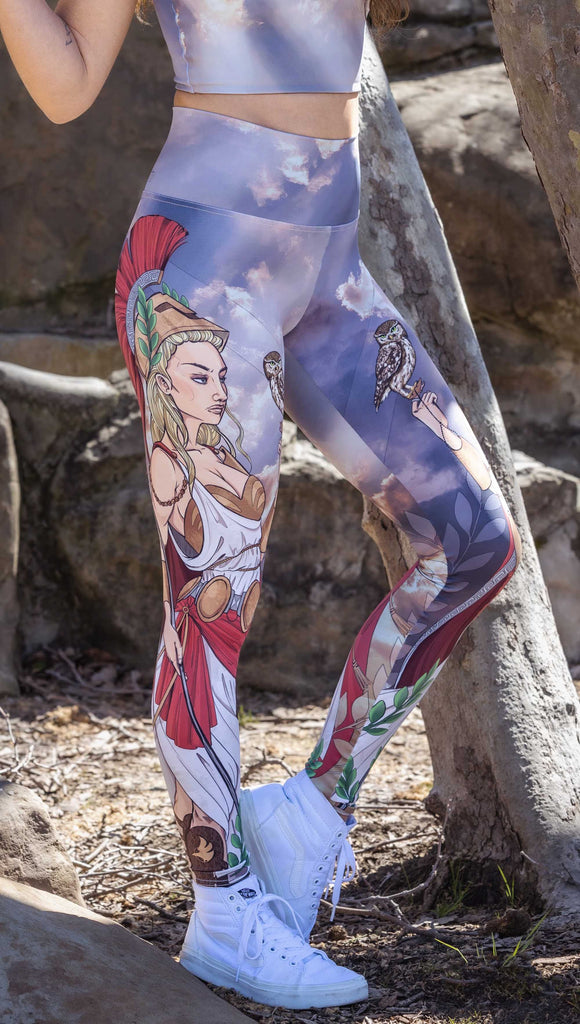 front side view of model wearing WERKSHOP Athena leggings. The leggings are printed with original artwork by Chriztina Marie and features Athena, the goddess of war standing on a cliff’s edge. She is holding a spear with one hand and her owl with the other. She is wearing a greek goddess dress/warrior hat with colors of cream, gold and red. Behind her is a beautiful cloudy sky with rays of light shining down onto her. The model is also wearing a matching top.