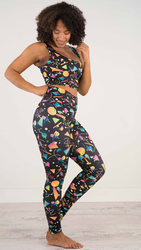Full body view of model wearing WERKSHOP white confetti athleisure leggings. The artwork on the leggings shows multi-colored circles, scribbles and triangles over a black background.