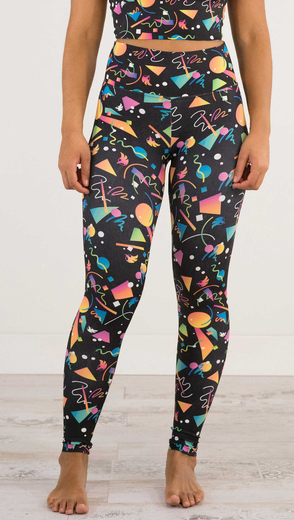 Waist down view of model wearing WERKSHOP white confetti athleisure leggings. The artwork on the leggings shows multi-colored circles, scribbles and triangles over a black background.